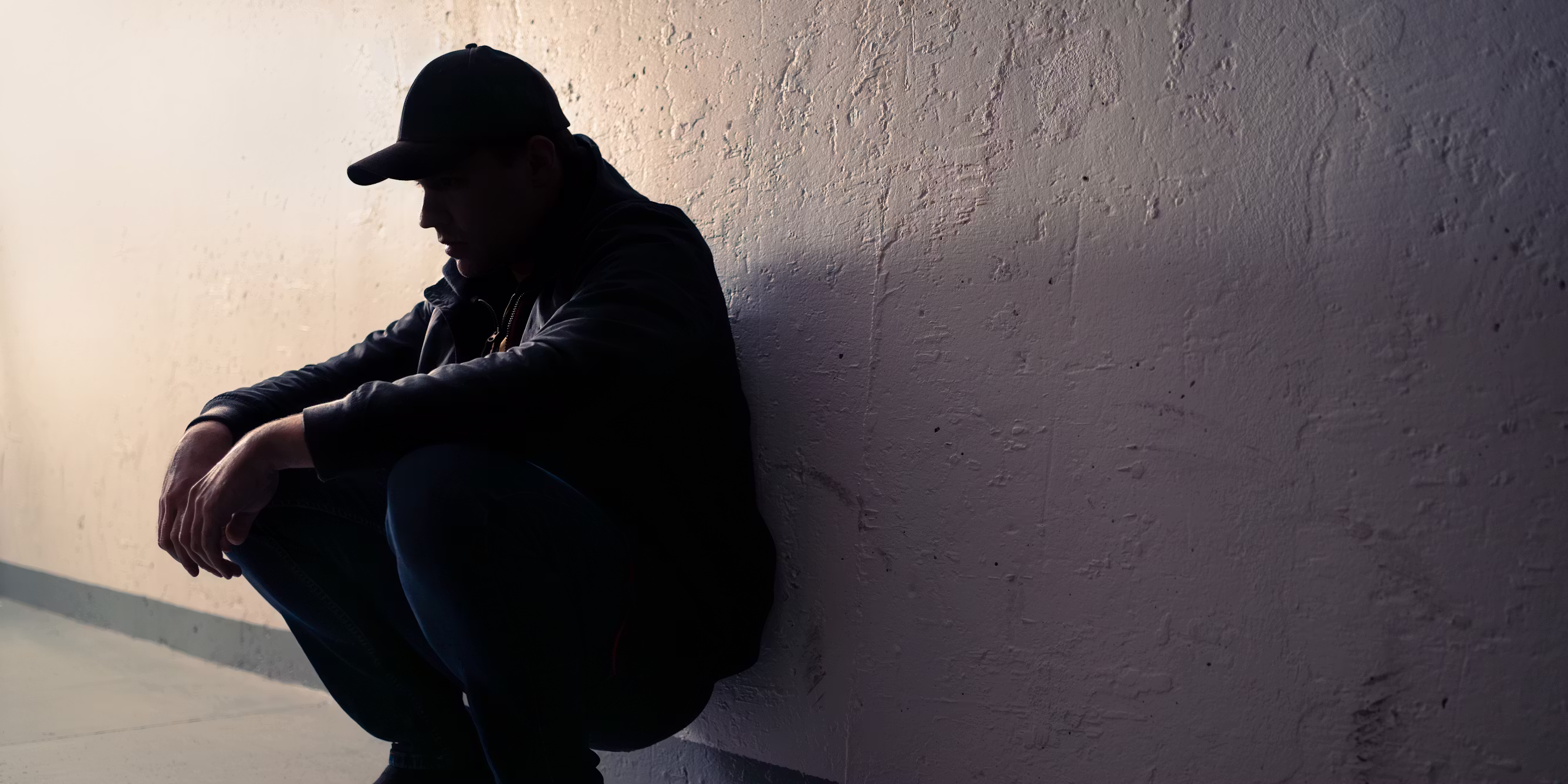 Photo of man crouched, leaning against wall, wearing a black baseball cap. He is shrouded in shadow.